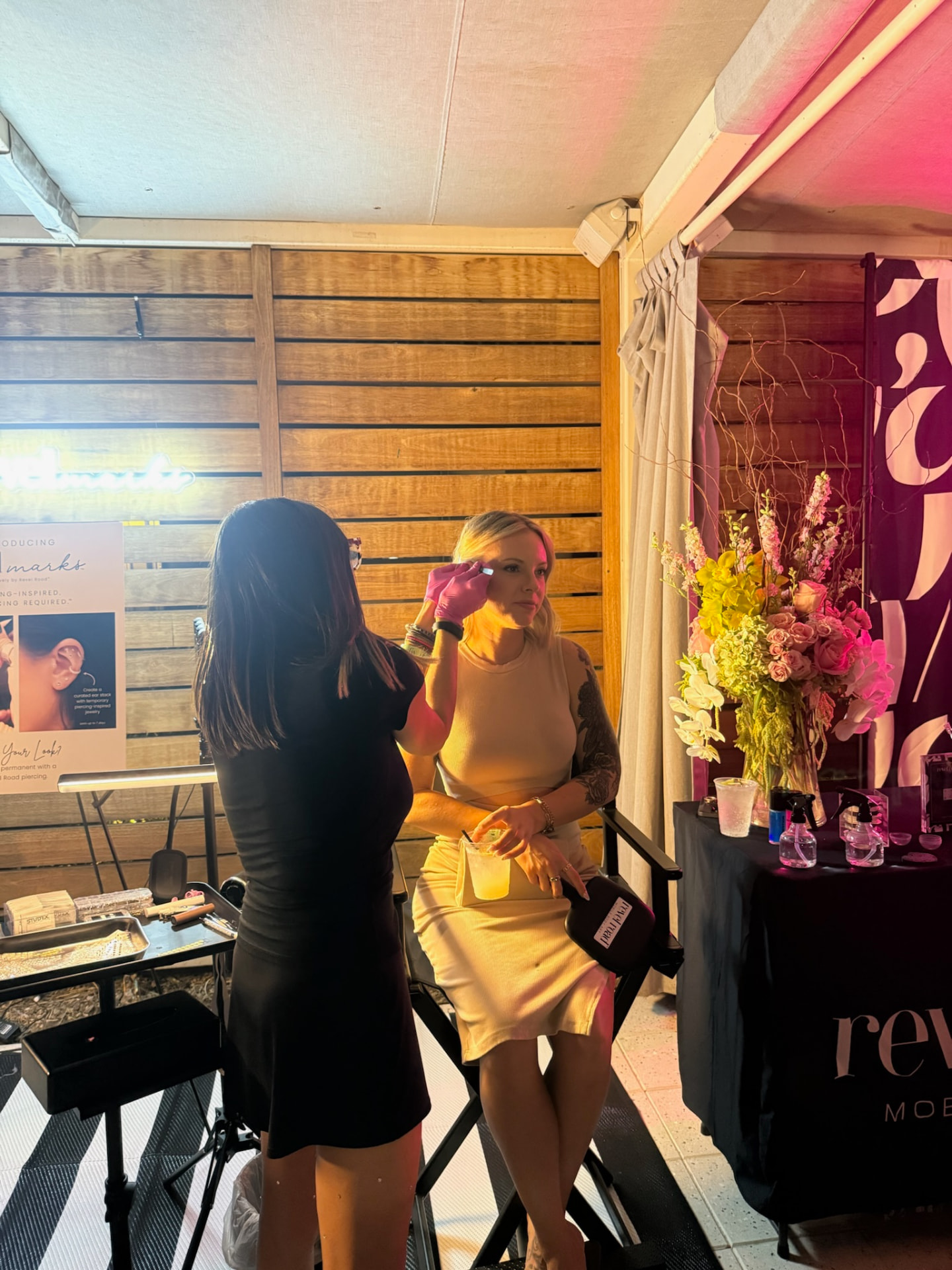 Pink-gloved Revel Road Artist adding crystal details to a seated guest beside the cabana flowers