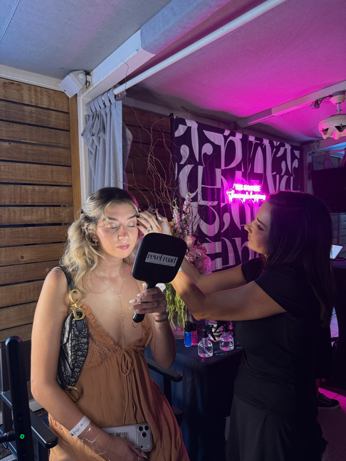 Revel Road Artist styling a guest holding the branded mirror beneath pink cabana lighting