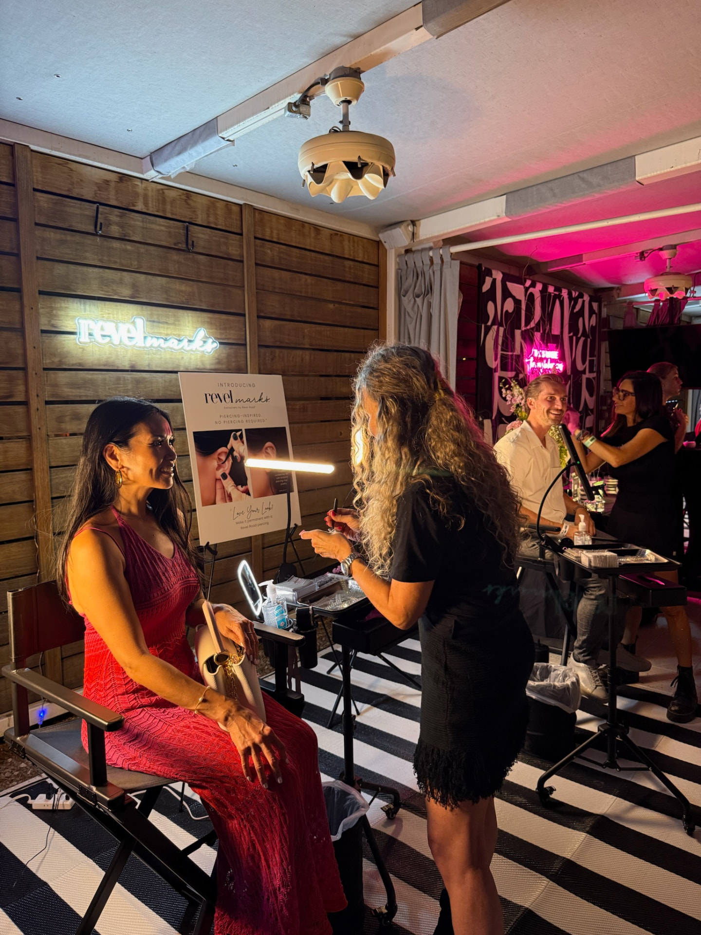 Two Revel Road Artists styling guests beneath the Revelmarks neon with branded backdrop and black-and-white rug